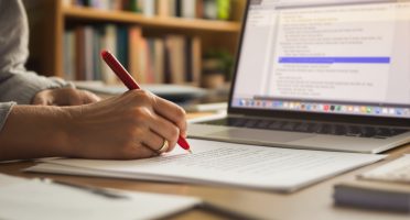 Close-up of an editor’s hand marking a manuscript with a red pen beside a laptop, symbolizing professional editing, developmental feedback, and copyediting services