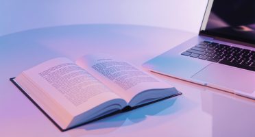 Open printed book beside a laptop on a clean, softly lit desk with blue and pink gradient lighting, representing professional book formatting and typesetting services.